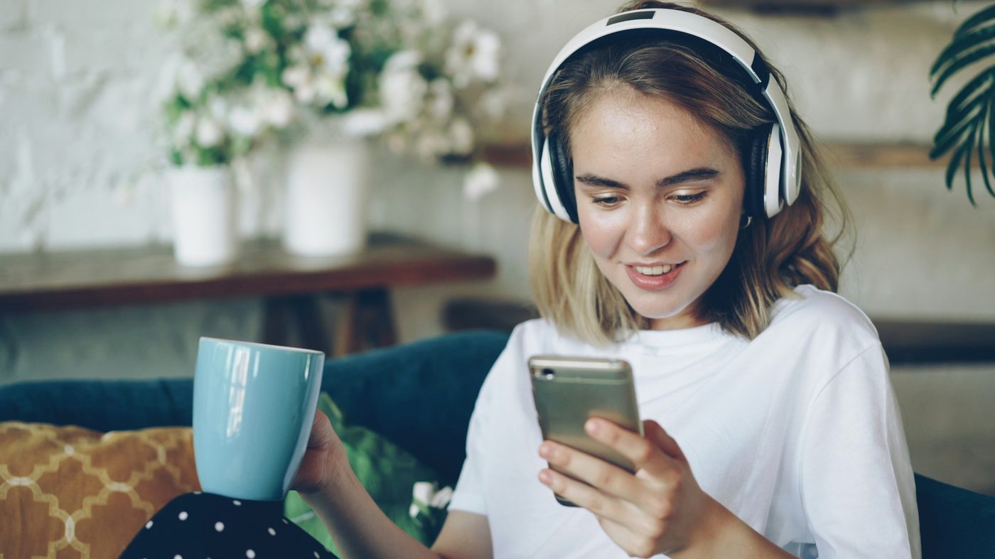 A woman in a cafe smiling at her phone, wearing white over-ear headphones, holding a blue mug