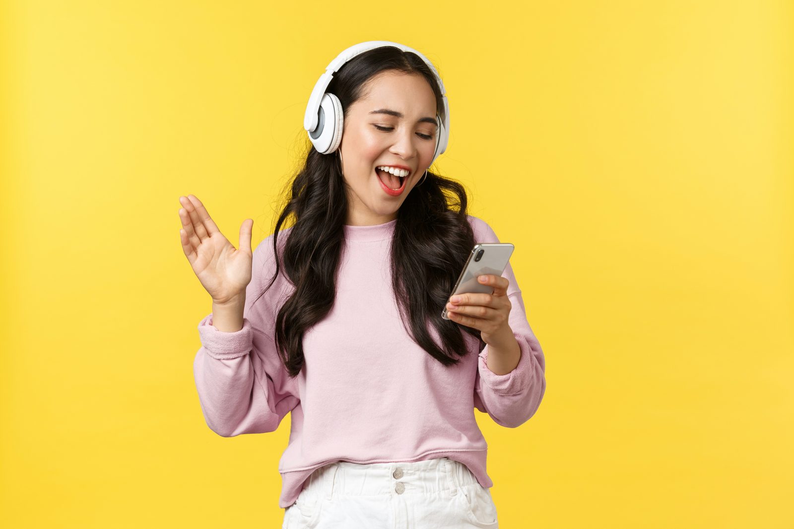 A woman in a pink sweater wearing white over-ear headphones, smiling and singing along to music while holding her phone