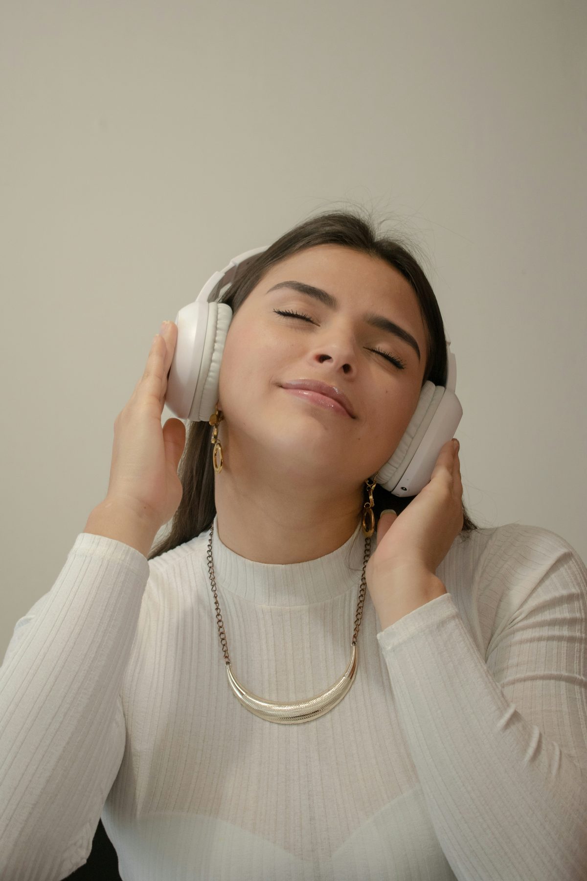 A woman with eyes closed wearing white over-ear headphones, hands gently cupped around them, lost in the music