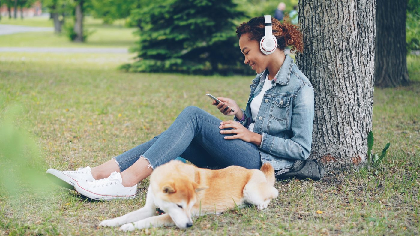 A woman sitting under a tree in a park with a dog, listening to music with white over-ear headphones